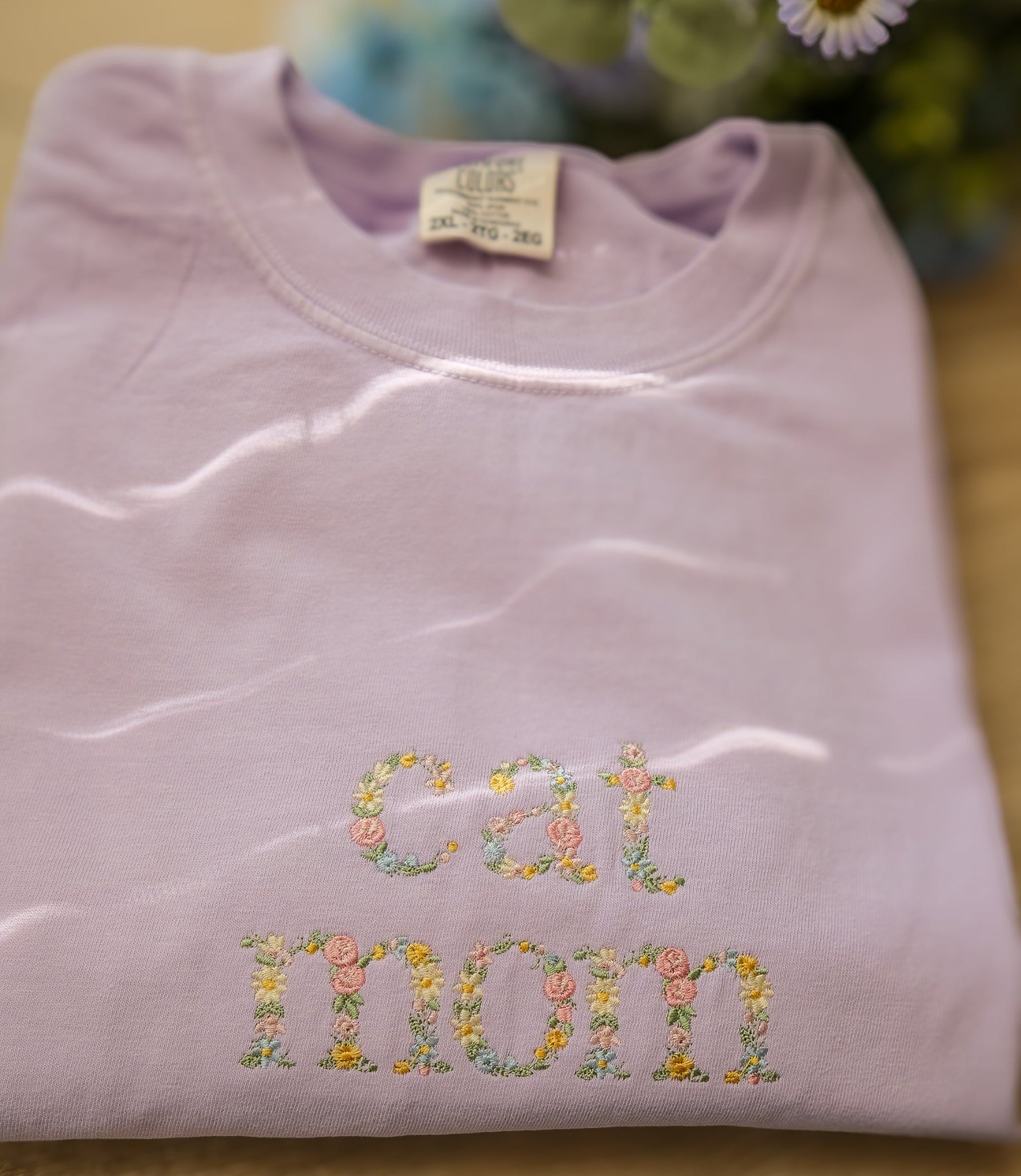 Folded orchid sweatshirt with embroidered floral text on a wooden surface with flowers in the background