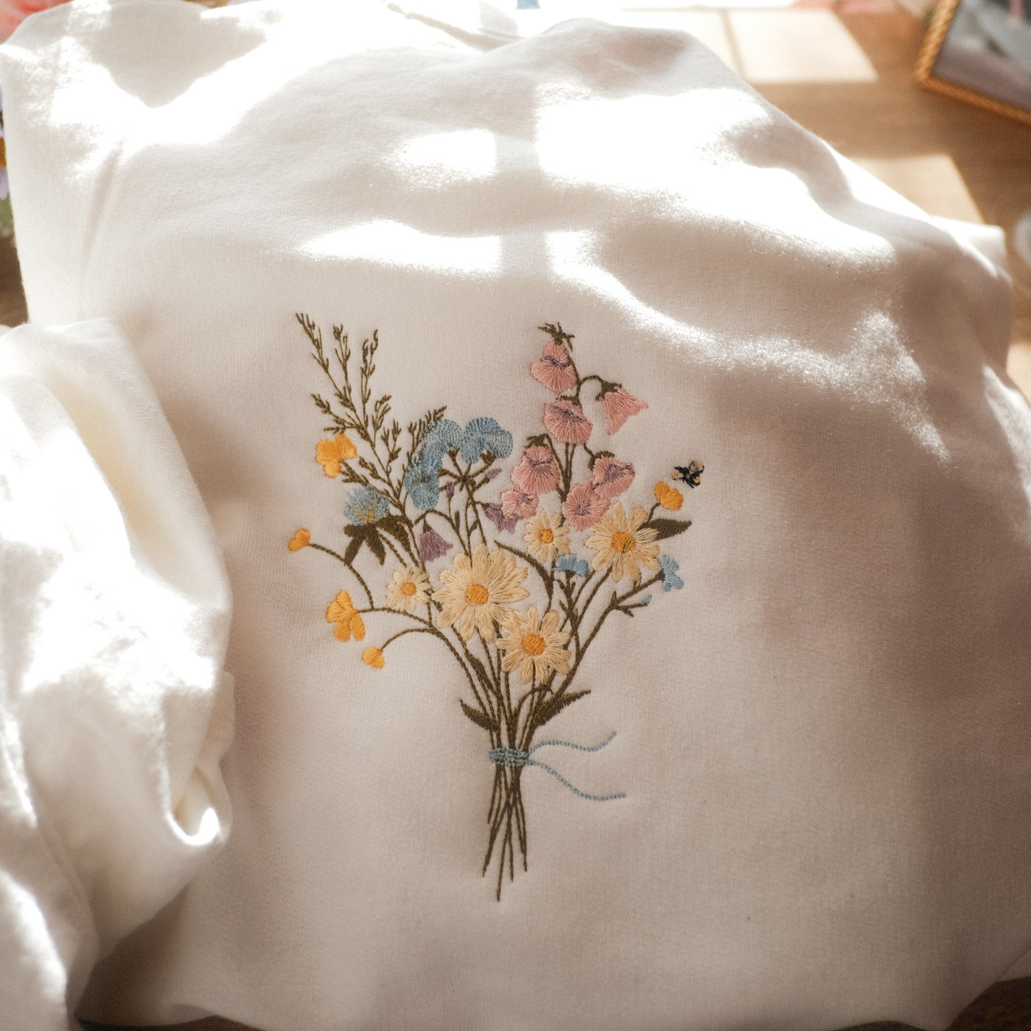 White sweatshirt with embroidered pastel wildflowers and a tiny bee detail, styled with soft floral decor and warm sunlight in the background