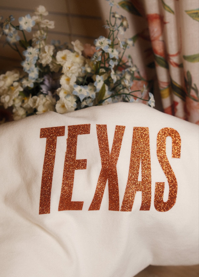 White T shirt with 'TEXAS' printed in orange glittery letters, placed on a surface with flowers and fabric in the background.