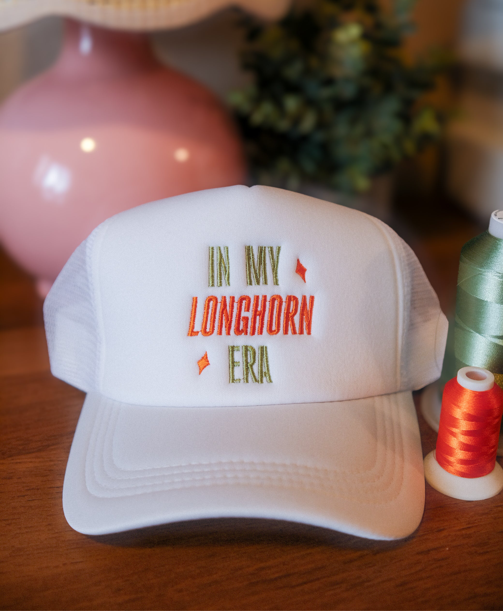 White cap with 'In My Longhorn Era' text on a wooden surface with thread spools.