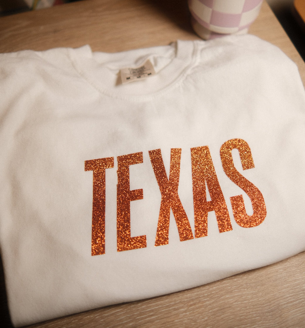 White shirt with 'TEXAS' printed in orange glitter on a wooden surface.
