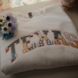 A folded white sweatshirt with the word "TEXAS" embroidered in colorful patchwork letters, featuring floral and gingham patterns. A small bouquet of flowers rests on top of the sweatshirt, adding a soft, cozy feel.