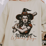 A person wearing a beige sweatshirt with a design featuring a fierce retro witch holding a bat with the text "Support Women's Rage" below the illustration. The witch is depicted with an intense expression, surrounded by small bats.