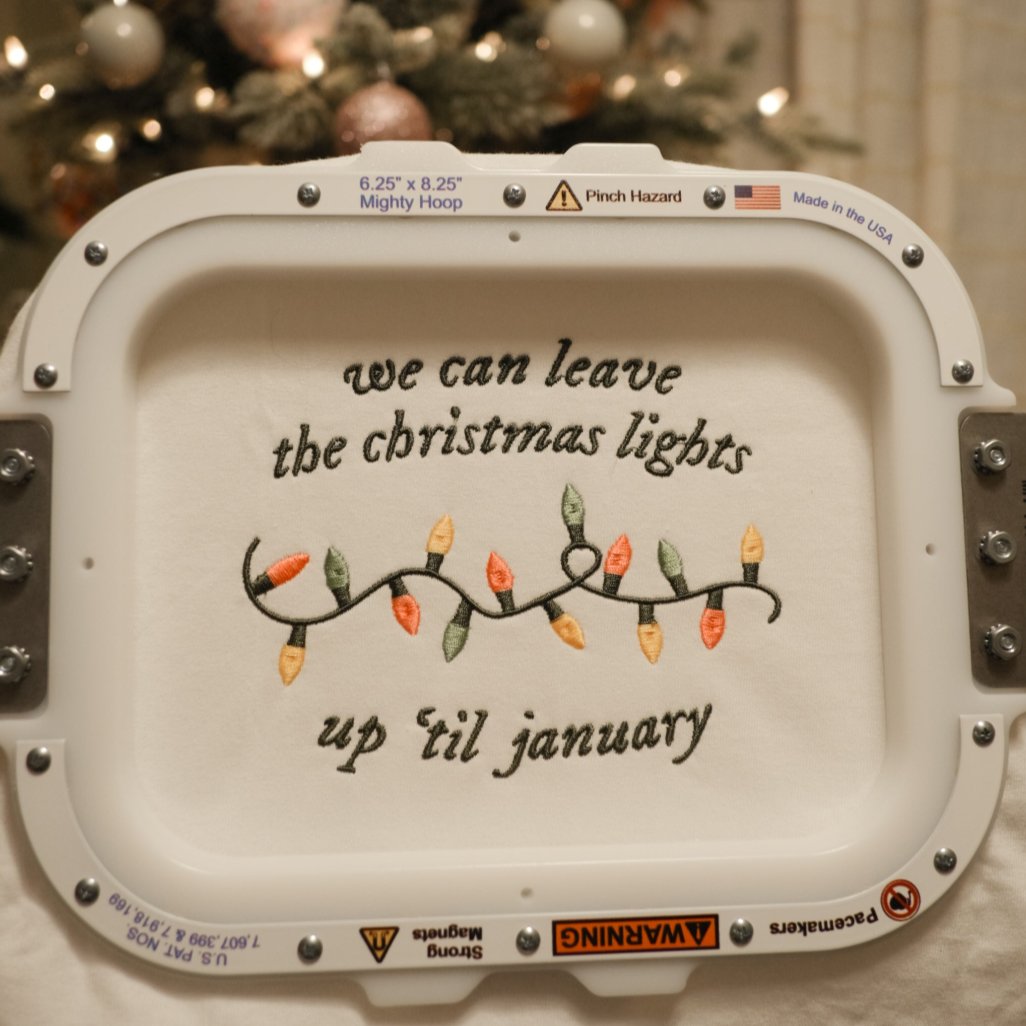 An embroidery hoop showcasing a festive design with the quote “We can leave the Christmas lights up ’til January” and a colorful embroidered string of lights, placed in front of a beautifully decorated Christmas tree.