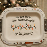An embroidery hoop showcasing a festive design with the quote “We can leave the Christmas lights up ’til January” and a colorful embroidered string of lights, placed in front of a beautifully decorated Christmas tree.