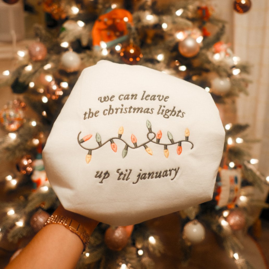 An embroidery hoop showcasing a festive design with the quote “We can leave the Christmas lights up ’til January” and a colorful embroidered string of lights, placed in front of a beautifully decorated Christmas tree.