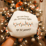 An embroidery hoop showcasing a festive design with the quote “We can leave the Christmas lights up ’til January” and a colorful embroidered string of lights, placed in front of a beautifully decorated Christmas tree.