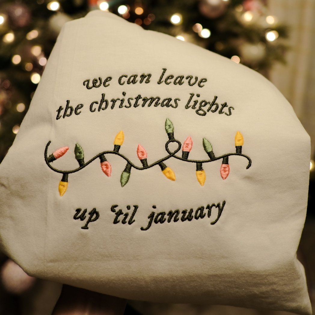 An embroidery hoop showcasing a festive design with the quote “We can leave the Christmas lights up ’til January” and a colorful embroidered string of lights, placed in front of a beautifully decorated Christmas tree.