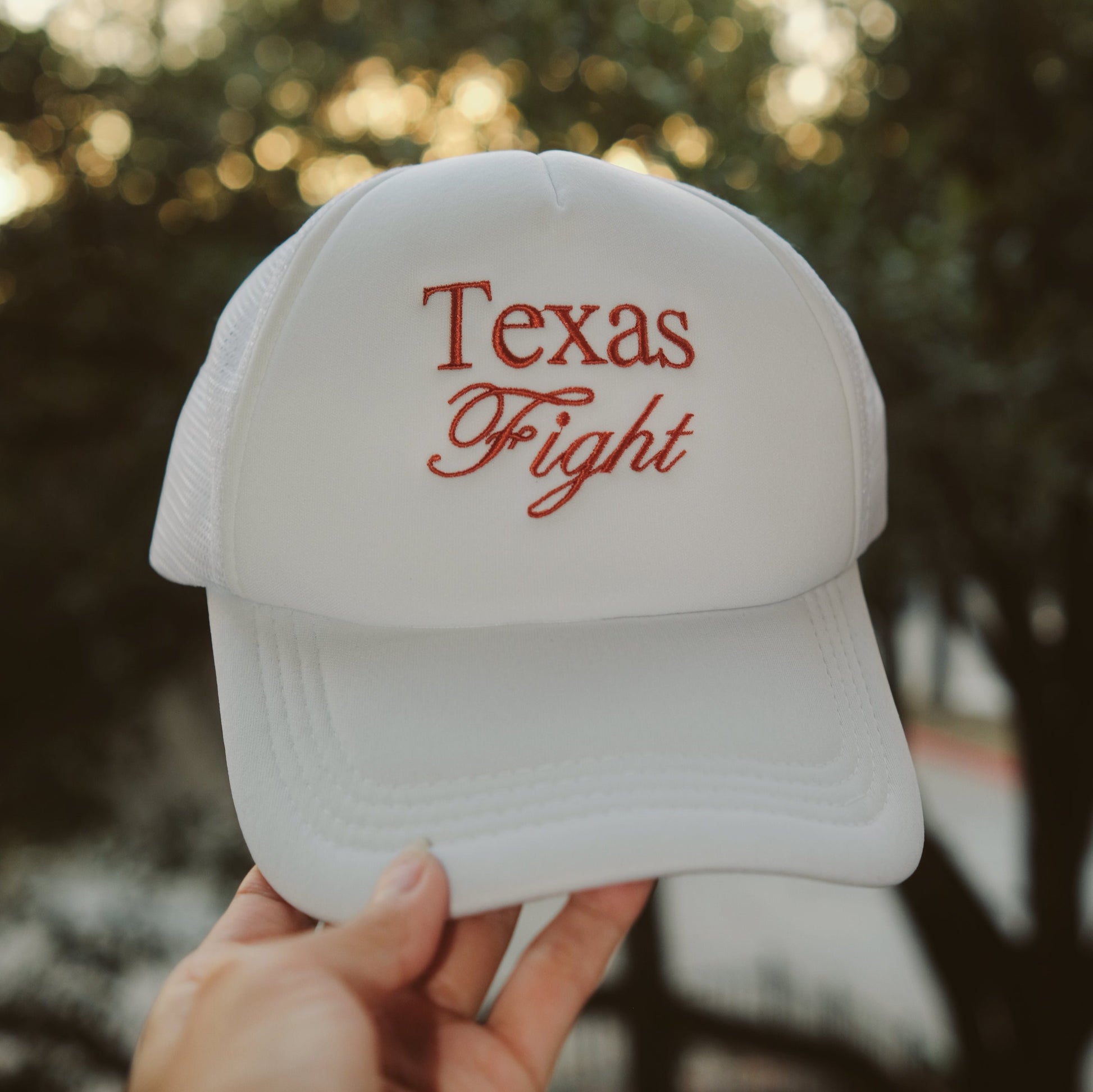 A hand holding a white trucker hat with the words "Texas Fight" embroidered in burnt orange script on the front, with a blurred background of trees and sunlight.