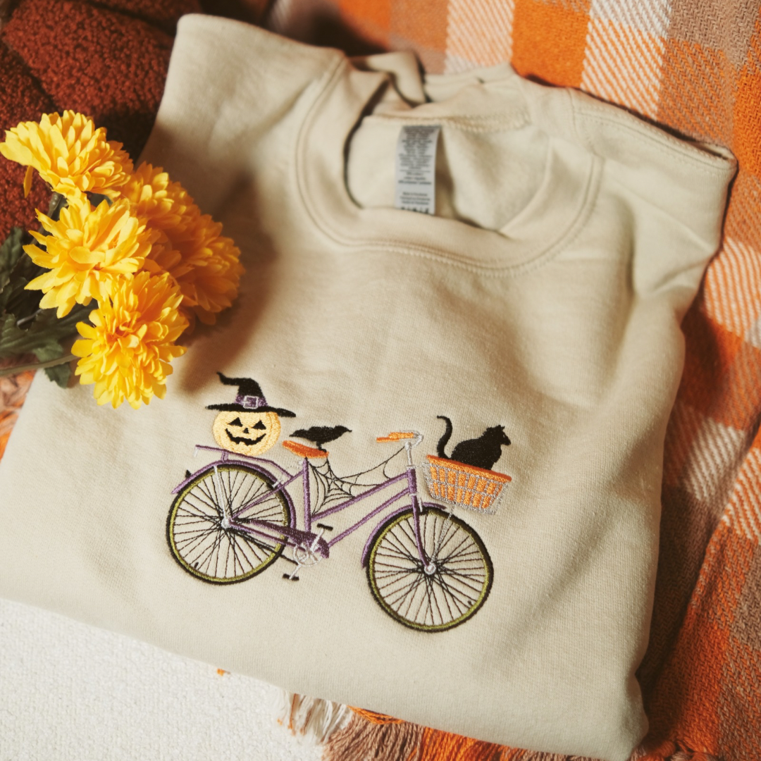 A folded beige sweatshirt with a detailed embroidery design featuring a purple bicycle decorated with a jack-o-lantern wearing a witch’s hat, a black cat in the basket, and a crow sitting on the bike. The sweatshirt is styled with yellow flowers and a cozy fall-themed blanket in the background.