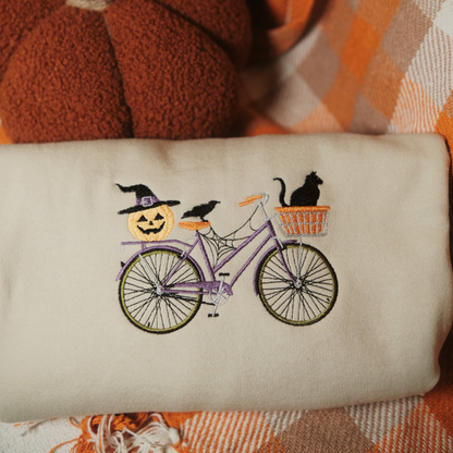 A folded beige sweatshirt with a detailed embroidery design featuring a purple bicycle decorated with a jack-o-lantern wearing a witch’s hat, a black cat in the basket, and a crow sitting on the bike. The sweatshirt is styled with yellow flowers and a cozy fall-themed blanket in the background.