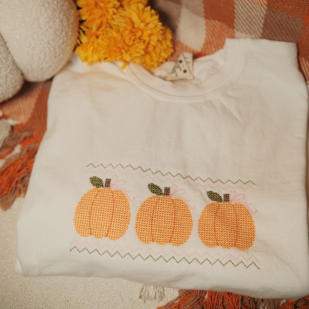 A folded white sweatshirt with an embroidered design of three orange pumpkins, featuring zigzag stitching above and below. Styled with yellow flowers and a cozy fall-themed blanket, perfect for autumn decor and vibes.