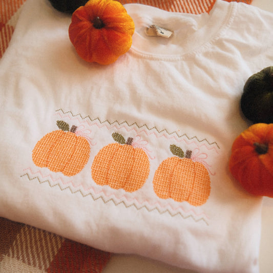 A folded white sweatshirt with an embroidered design of three orange pumpkins, featuring zigzag stitching above and below. Styled with yellow flowers and a cozy fall-themed blanket, perfect for autumn decor and vibes.