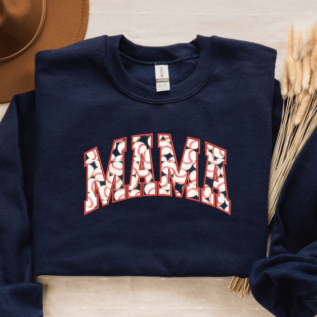 Navy sweatshirt with 'MAMA' varsity text filled with baseball-themed fabric, styled with a brown hat and wheat stems on a neutral background.