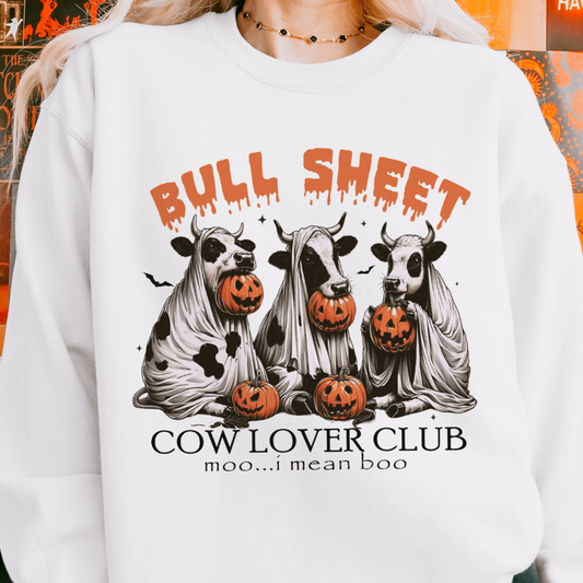 A person wearing a white sweatshirt with a design featuring three cows dressed as ghosts holding jack-o'-lanterns, with the text "Bull Sheet Cow Lover Club" above and "moo...I mean boo" below the illustration. The background is decorated with Halloween-themed images.