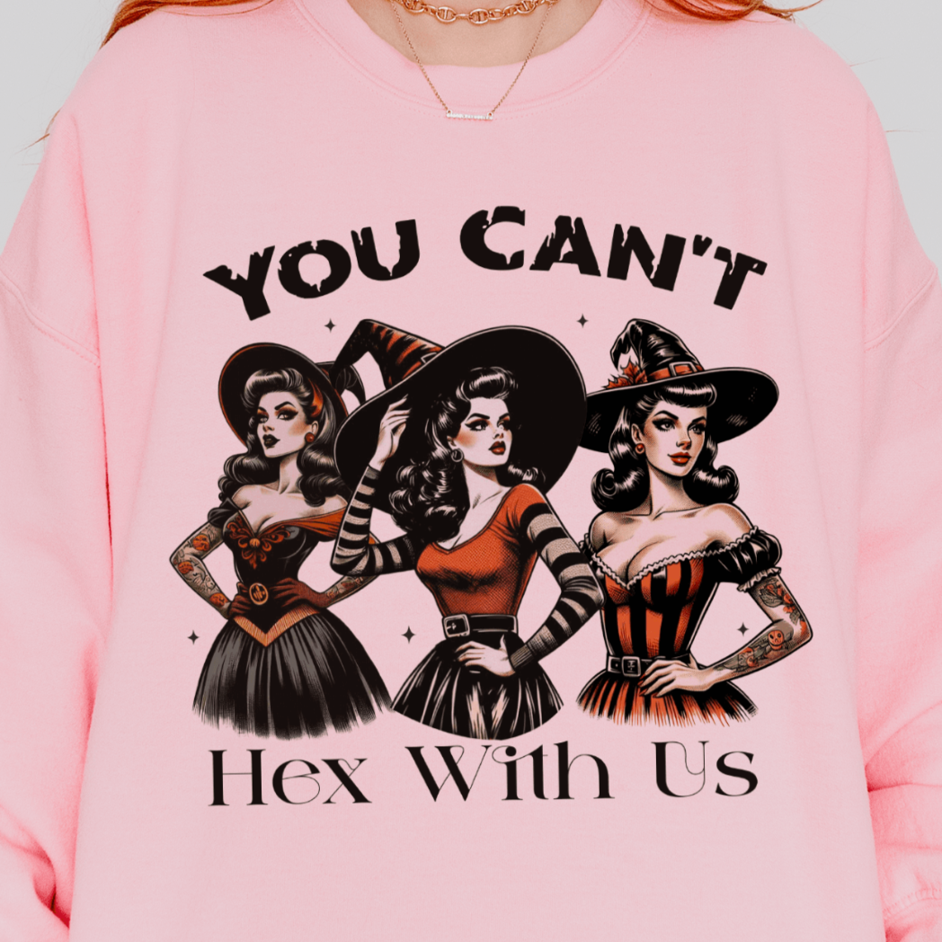 A person wearing a pink sweatshirt with a design featuring three retro witches in fashionable outfits with the text "You Can't Hex With Us" above and below the illustration.