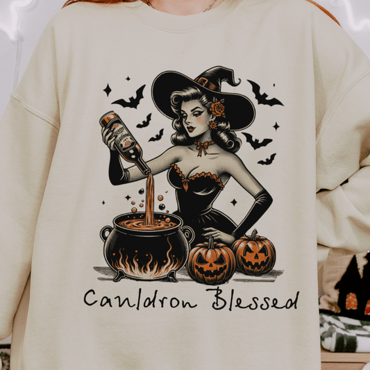 A person wearing a beige sweatshirt with a design featuring a retro witch pouring a potion into a cauldron, surrounded by pumpkins and bats, with the text "Cauldron Blessed" below the illustration. The background includes Halloween-themed decorations.