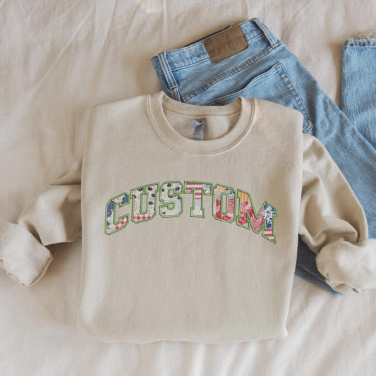 A folded beige sweatshirt with colorful patchwork letters spelling "CUSTOM," showcasing a personalized design option. Styled with jeans on a neutral background for a casual, cozy vibe.