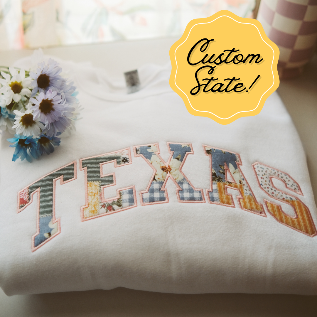 A folded white sweatshirt with "Texas" embroidered in colorful patchwork letters, featuring floral and gingham patterns. Customizable state design with a soft, cozy vibe, accompanied by a bouquet of flowers in the background.