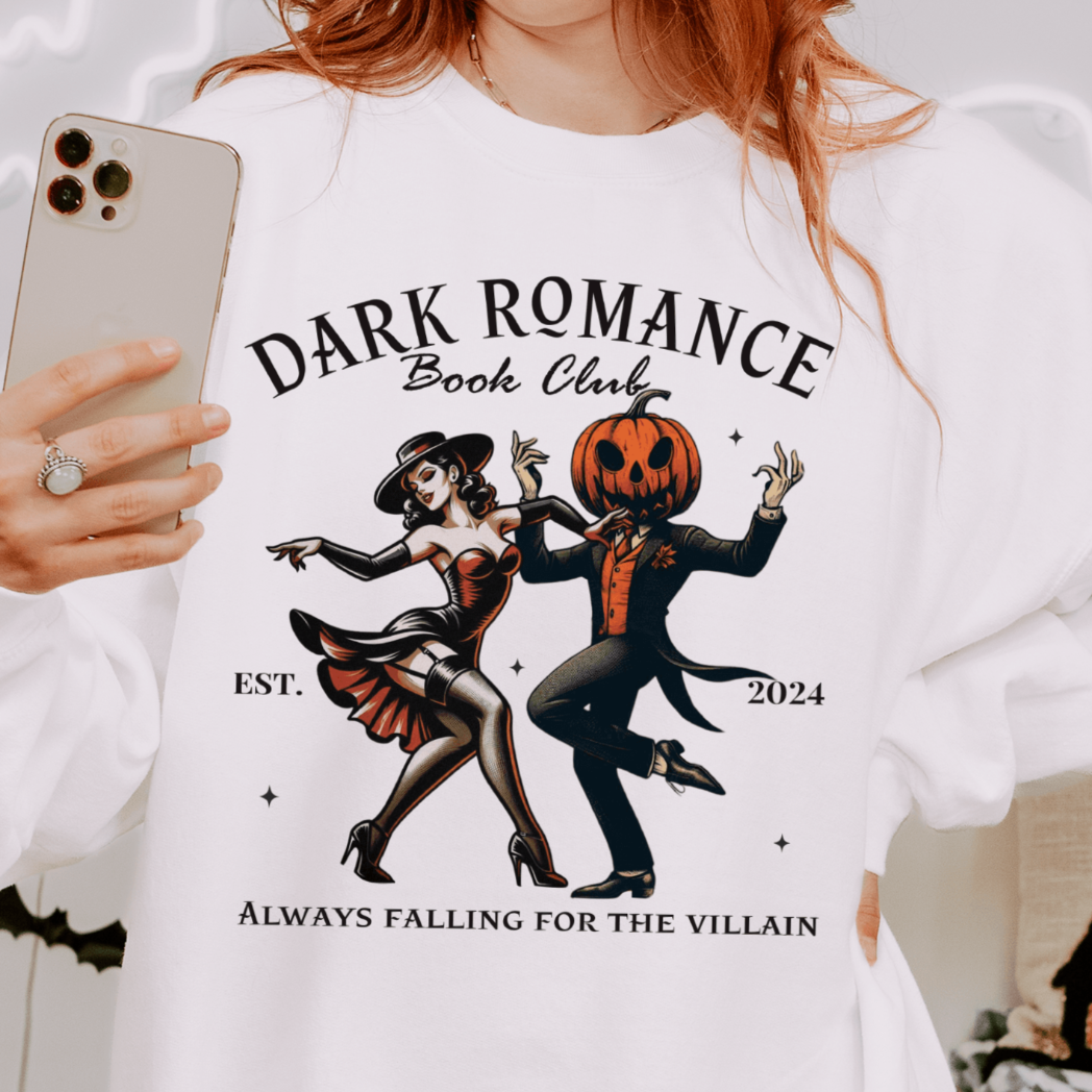 A person wearing a white sweatshirt with a design featuring a retro witch dancing with a pumpkin-headed man in a suit, with the text "Dark Romance Book Club - Est. 2024 - Always Falling for the Villain" above and below the illustration. The background includes Halloween-themed decorations.