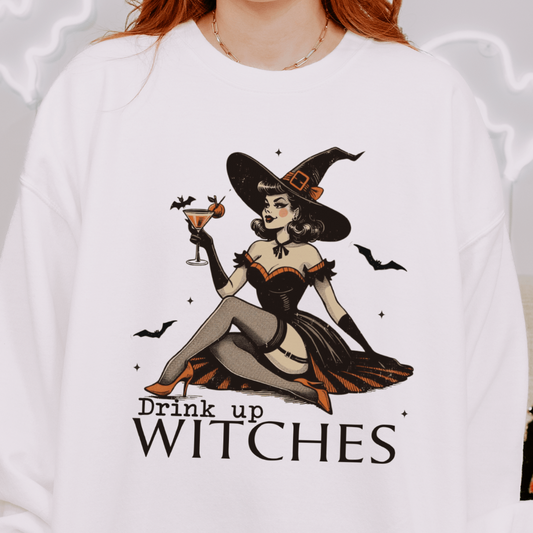 A person wearing a white sweatshirt with a design featuring a retro witch sitting down and holding a cocktail, with the text "Drink Up Witches" below the illustration. The background includes Halloween-themed decorations.