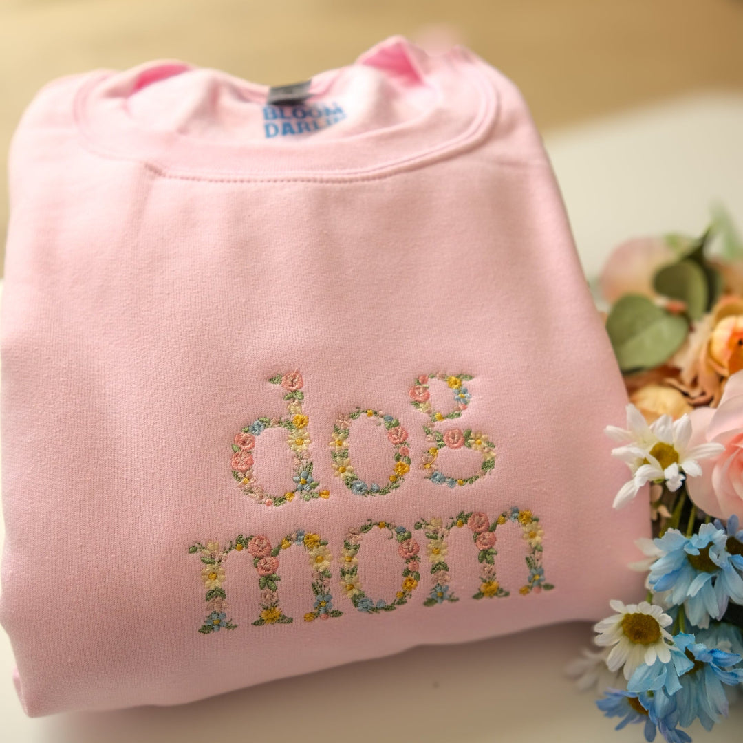 Pink sweatshirt with 'dog mom' text embroidered in colorful letters on a light surface with flowers.
