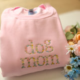 Pink sweatshirt with 'dog mom' text embroidered in colorful letters on a light surface with flowers.