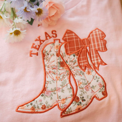 A folded pink t-shirt featuring embroidered floral cowboy boots with a plaid bow and the word "Texas" above. Styled with flowers on the side, perfect for personalized state pride apparel.