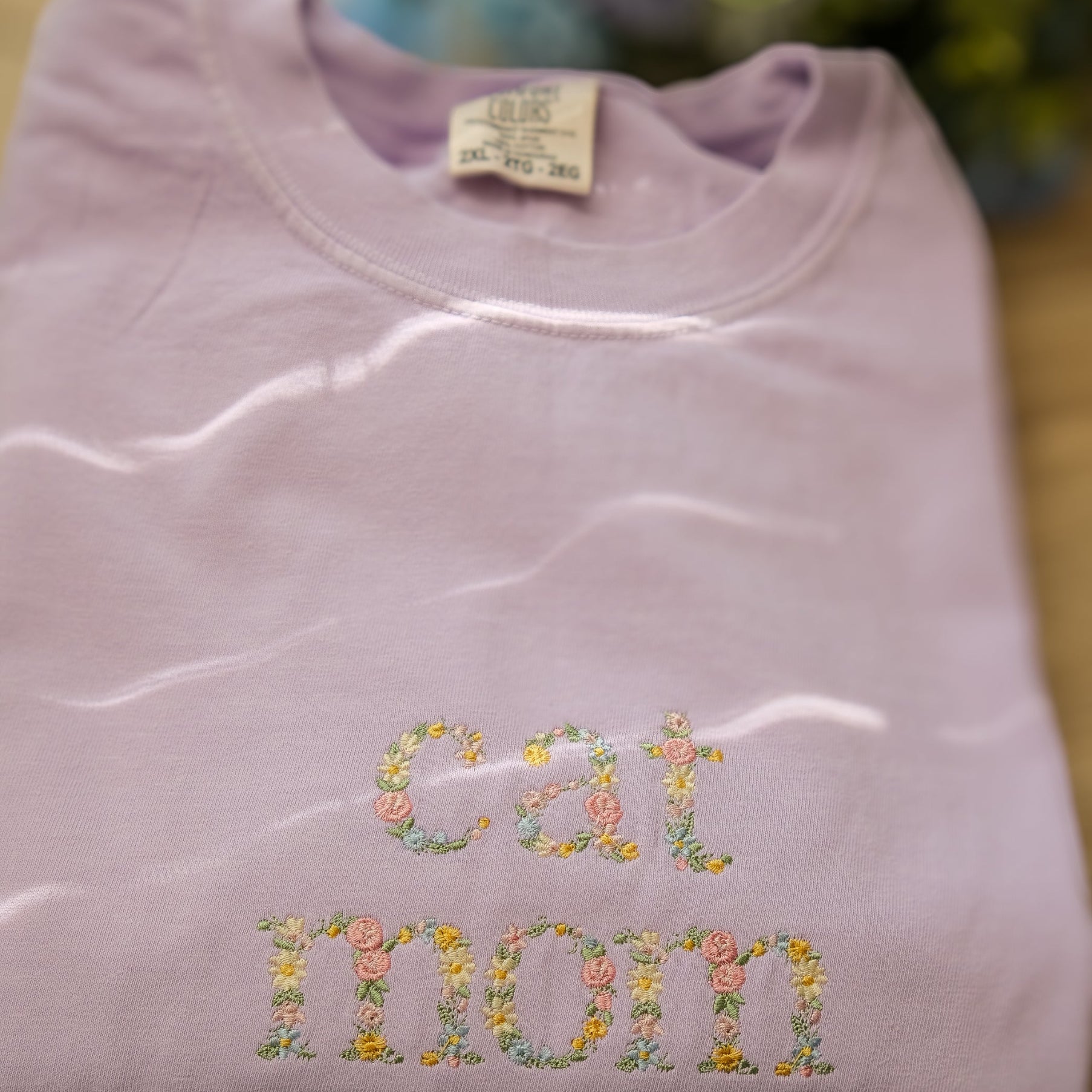 Folded orchid sweatshirt with embroidered floral text on a wooden surface with flowers in the background