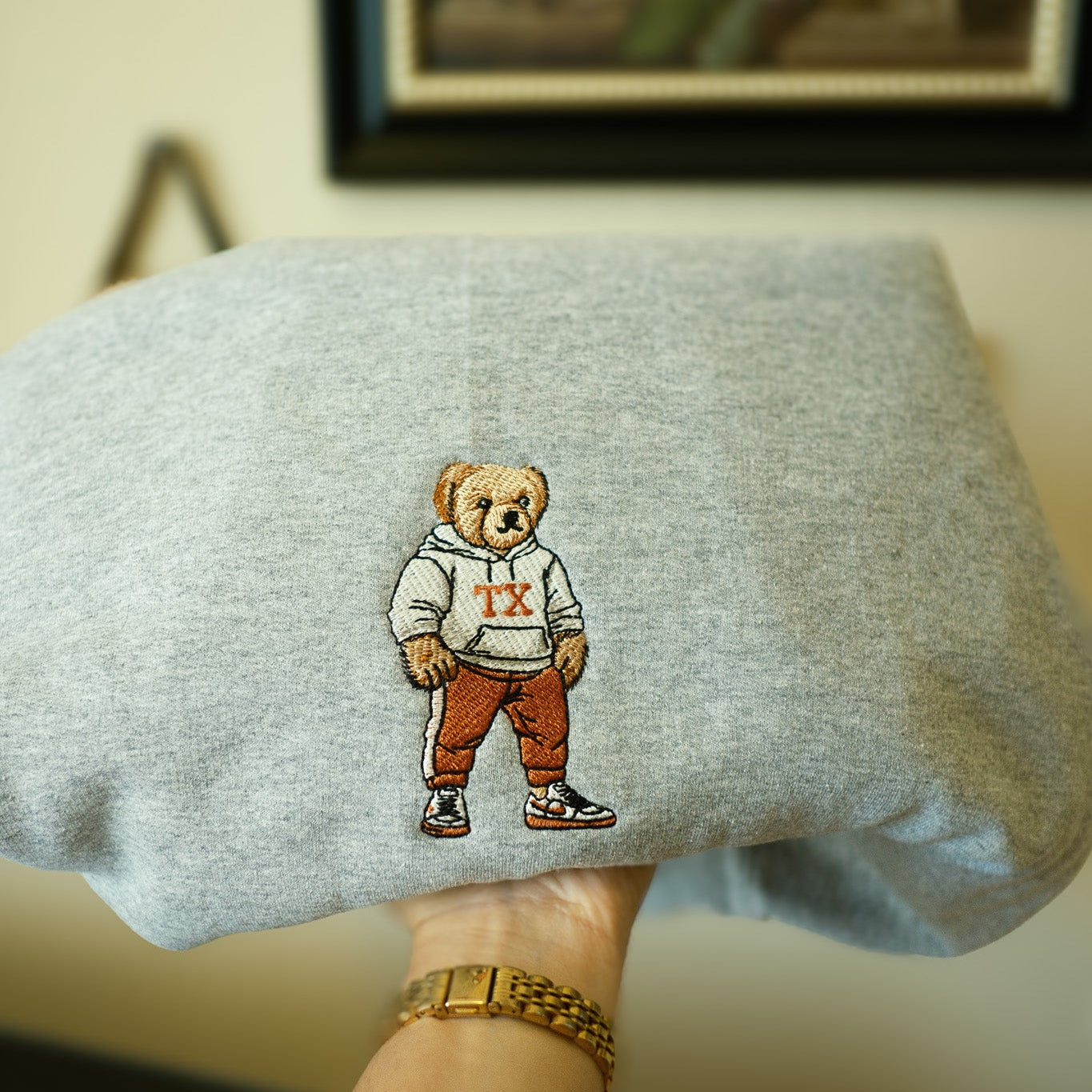 Gray sweatshirt with embroidered UT Austin bear design held by a person wearing a gold watch.