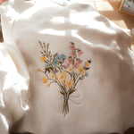 White sweatshirt with embroidered pastel wildflowers and a tiny bee detail, styled with soft floral decor and warm sunlight in the background