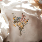 White sweatshirt with embroidered pastel wildflowers and a tiny bee detail, styled with soft floral decor and warm sunlight in the background