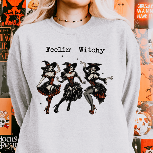 A person wearing a grey sweatshirt with a design featuring three retro witches in different stylish poses with the text "Feelin' Witchy" above the illustration. The background includes Halloween-themed decorations.