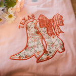 A folded pink t-shirt featuring embroidered floral cowboy boots with a plaid bow and the word "Texas" above. Styled with flowers on the side, perfect for personalized state pride apparel.