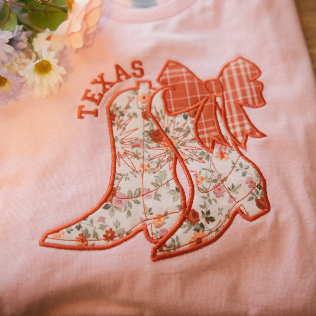 A folded pink t-shirt featuring embroidered floral cowboy boots with a plaid bow and the word "Texas" above. Styled with flowers on the side, perfect for personalized state pride apparel.