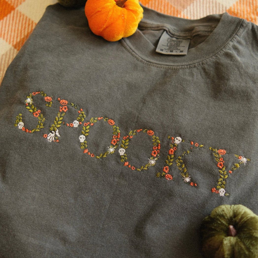 A folded dark grey t-shirt with the word "SPOOKY" embroidered in floral patterns with skulls and spiders. Styled with small pumpkins and an autumn blanket, perfect for cozy fall and Halloween vibes.