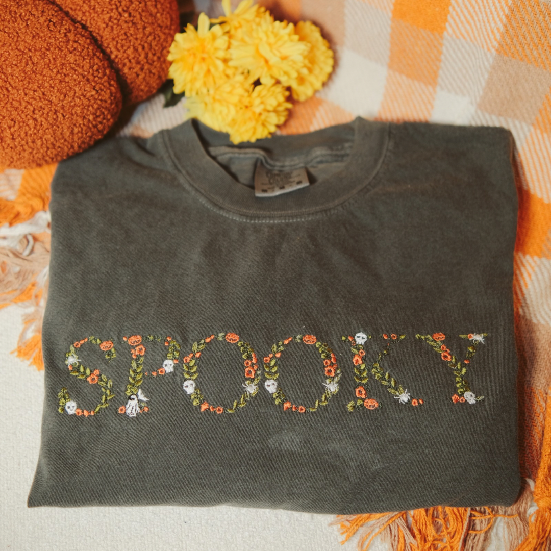 A folded dark grey t-shirt with the word "SPOOKY" embroidered in floral patterns with skulls and spiders. Styled with small pumpkins and an autumn blanket, perfect for cozy fall and Halloween vibes.