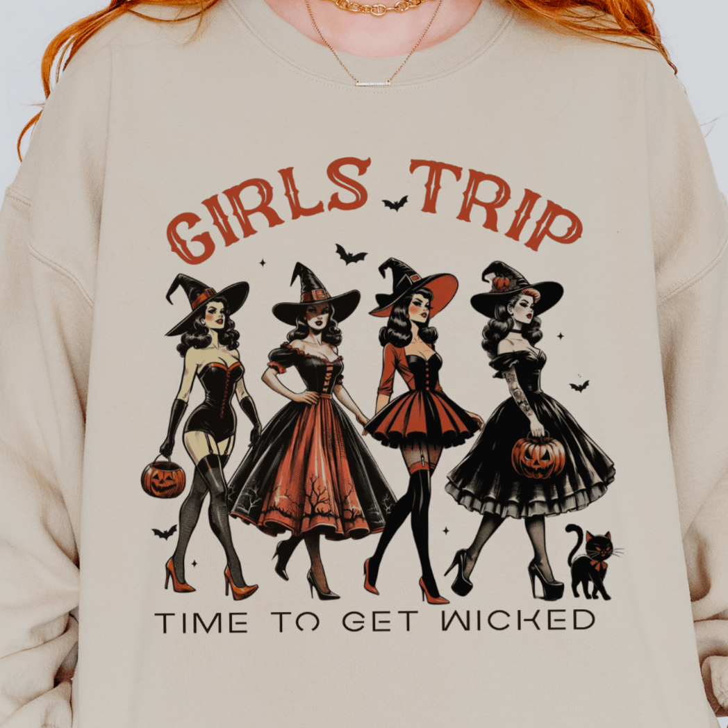 A person wearing a beige sweatshirt with a design featuring four stylish witches in different retro outfits, each holding a Halloween-themed item, with the text "Girls Trip - Time to Get Wicked" above and below the illustration.