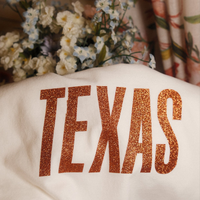 White T shirt with 'TEXAS' printed in orange glittery letters, placed on a surface with flowers and fabric in the background.