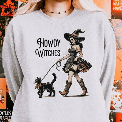 A person wearing a grey sweatshirt with a design featuring a retro witch in a Western-style outfit walking a black cat on a leash, with the text "Howdy Witches" beside the illustration.