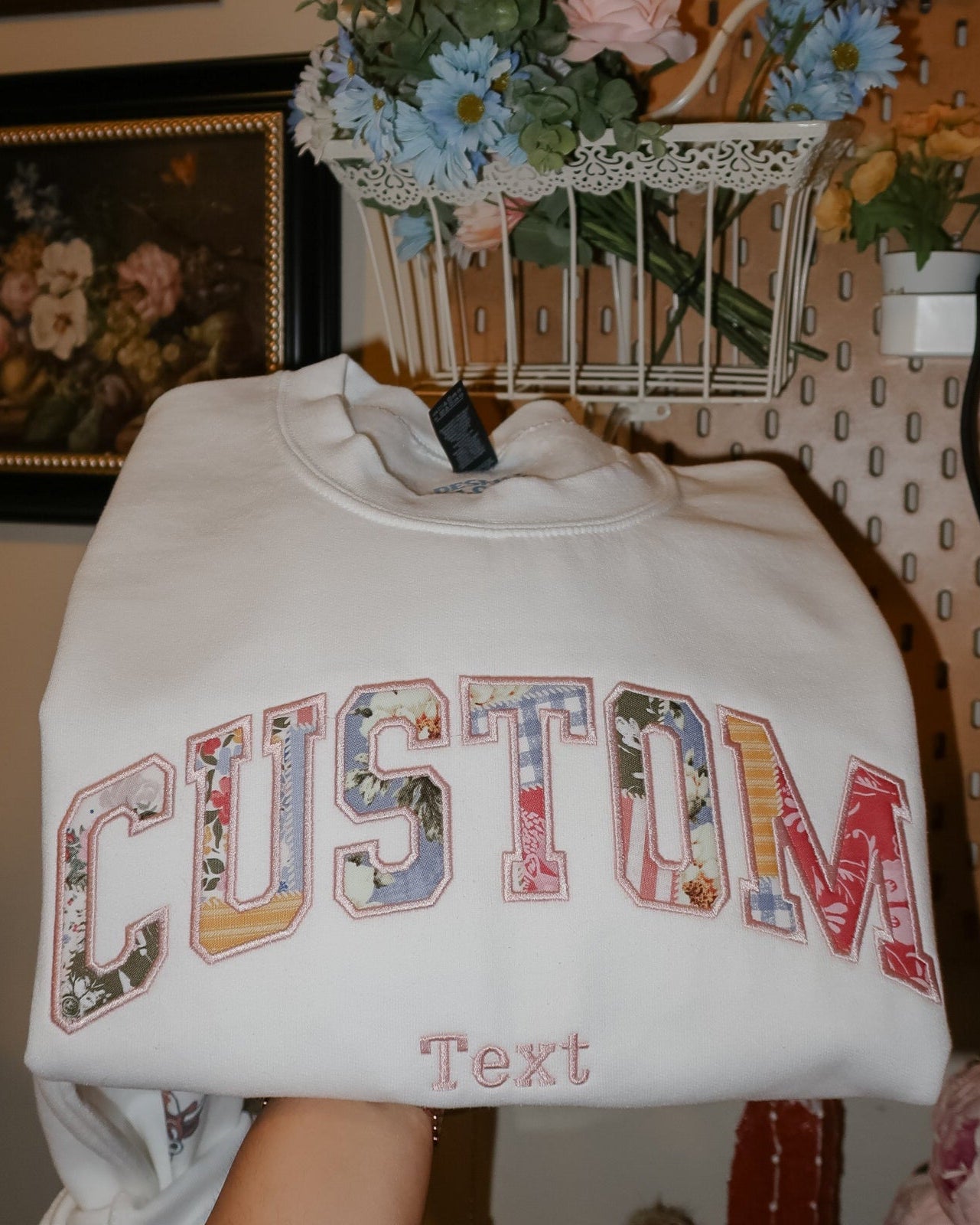 White sweatshirt with 'CUSTOM Text' printed on it, held in front of a decorative background with flowers and artwork.