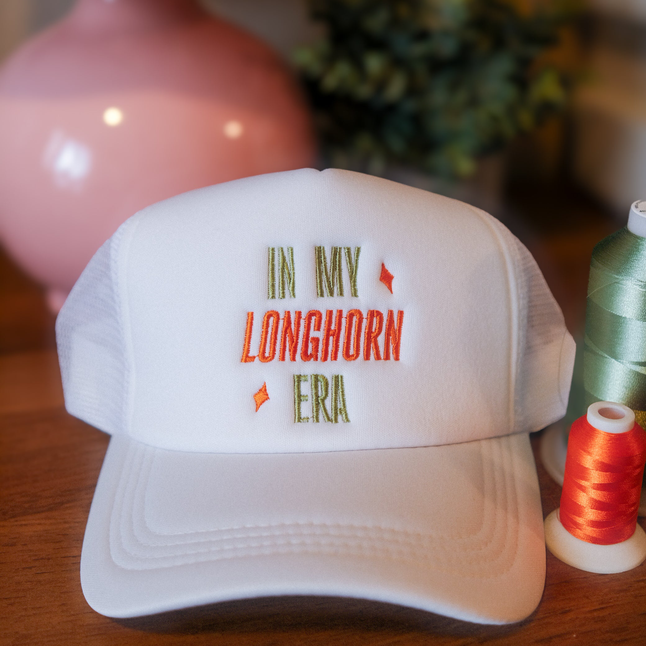 White cap with 'In My Longhorn Era' text on a wooden surface with thread spools.