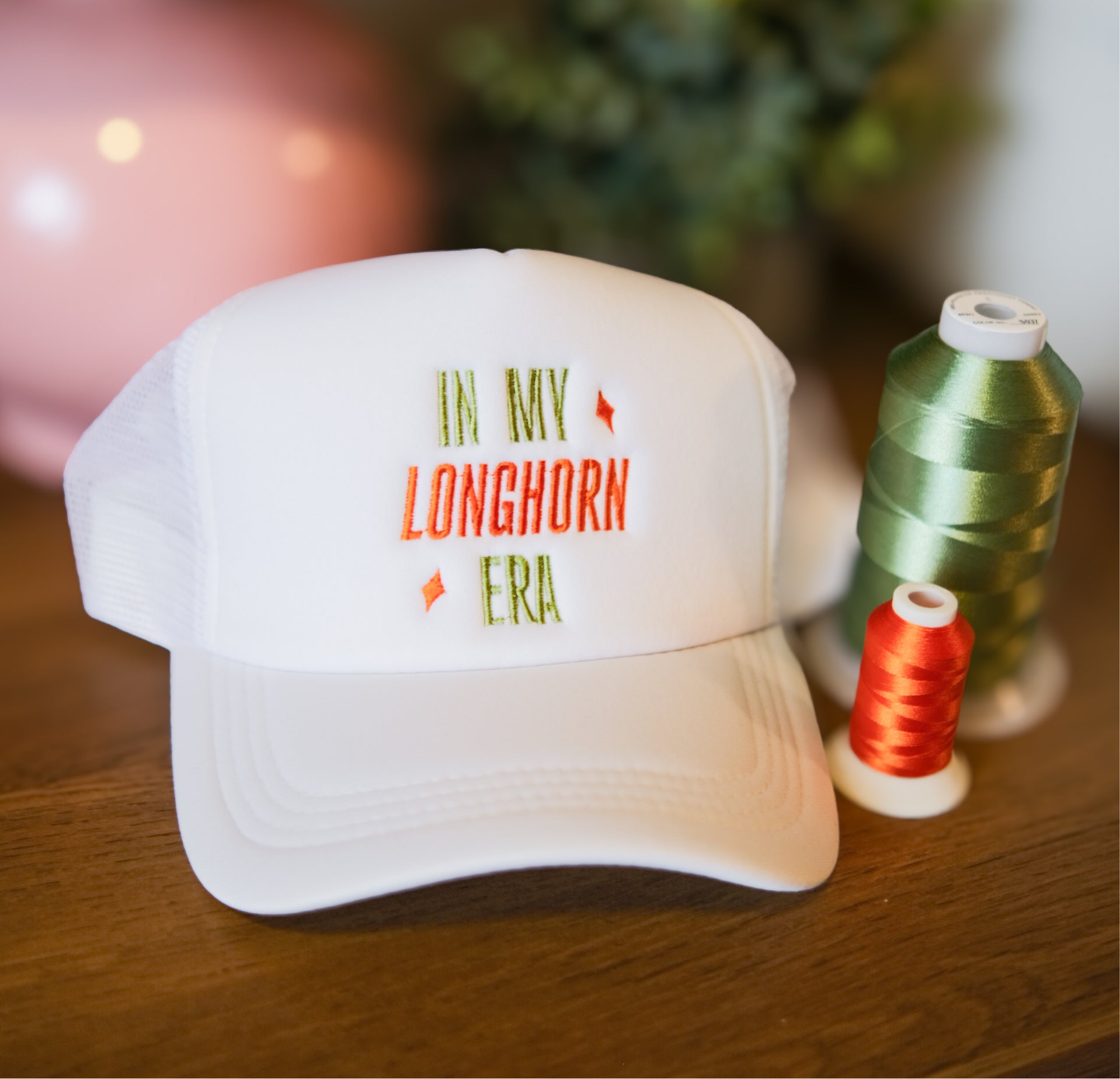 White cap with 'In My Longhorn Era' text on a wooden surface with spools of thread.