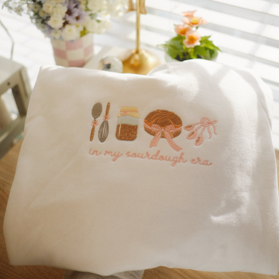 "White sweatshirt in embroidery hoop with 'In My Sourdough Era' design featuring a whisk, jar of starter, loaf of bread, and tied utensils in soft pink and brown tones.