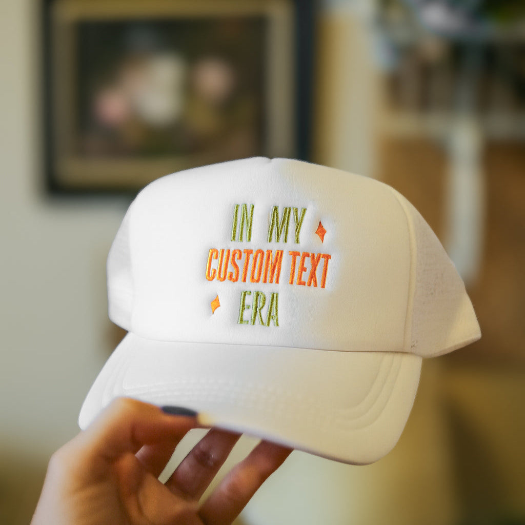 White cap with custom text held by a hand against a blurred indoor background