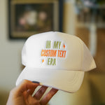White cap with custom text held by a hand against a blurred indoor background