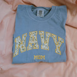 Folded blue t shirt with 'Navy Mom' text on a pink fabric background