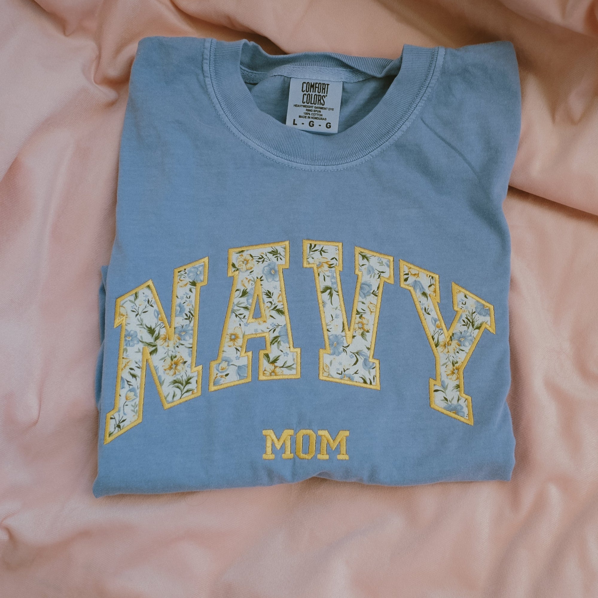 Folded blue t shirt with 'Navy Mom' text on a pink fabric background