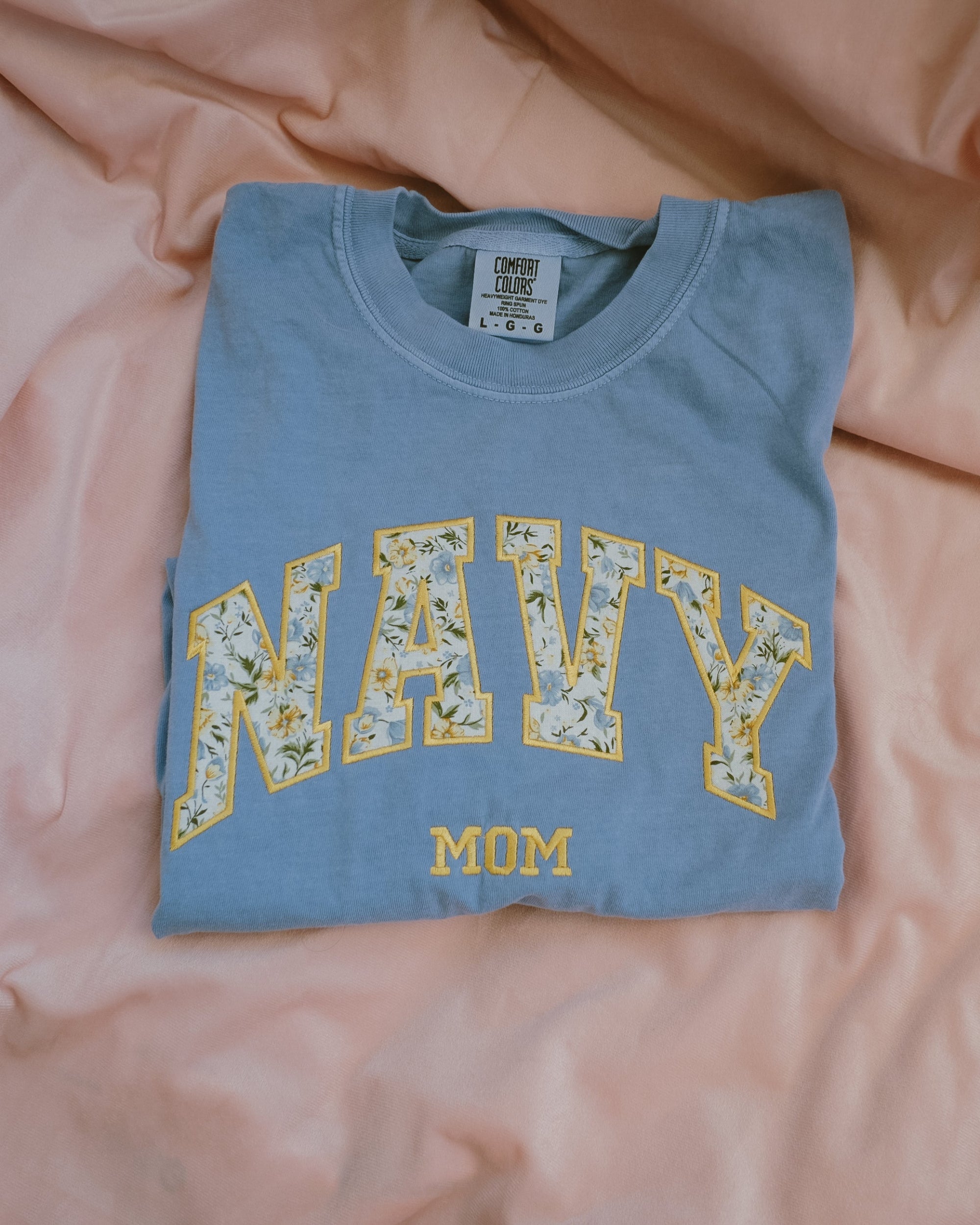 Folded blue t shirt with 'Navy Mom' text on a pink fabric background