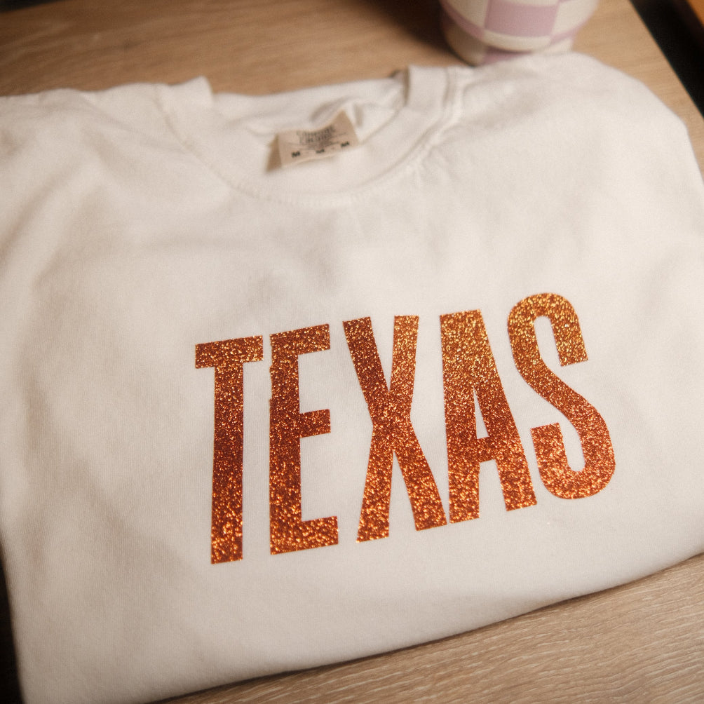 White shirt with 'TEXAS' printed in orange glitter on a wooden surface.